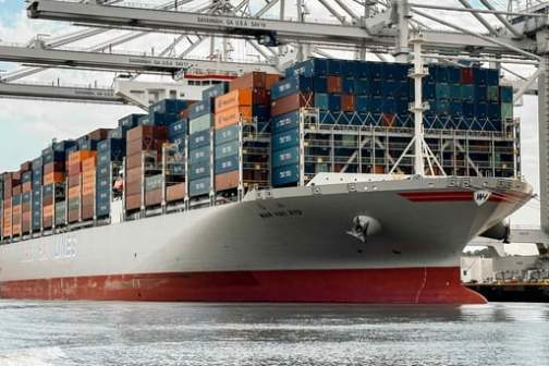 Large cargo ship loaded with colorful containers docked at a port with cranes above.