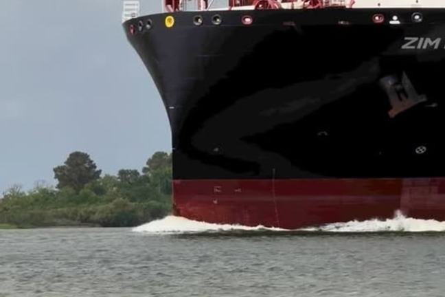 Large cargo ship with stacked containers on a river near greenery under cloudy sky.