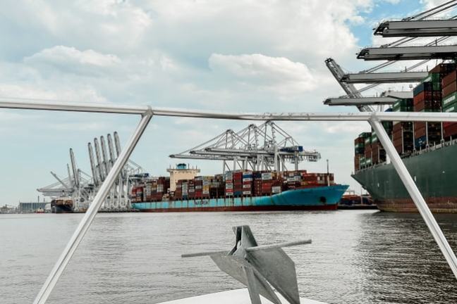 View from boat of cargo ships and cranes at a busy port under cloudy sky.