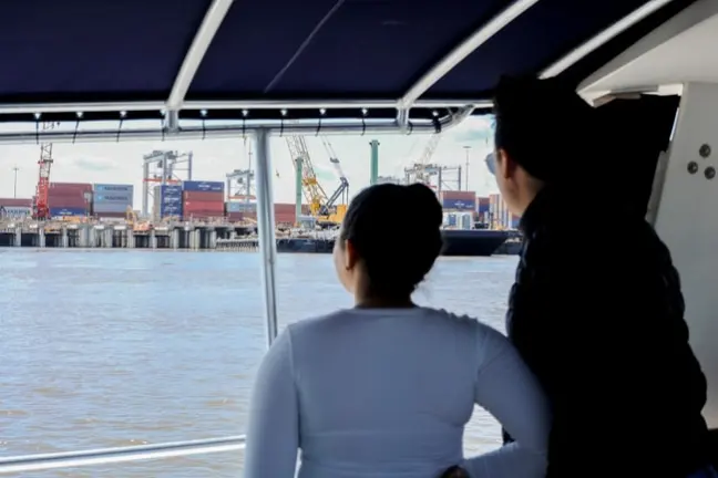 Two people on a boat looking at shipping containers and cranes across the water.